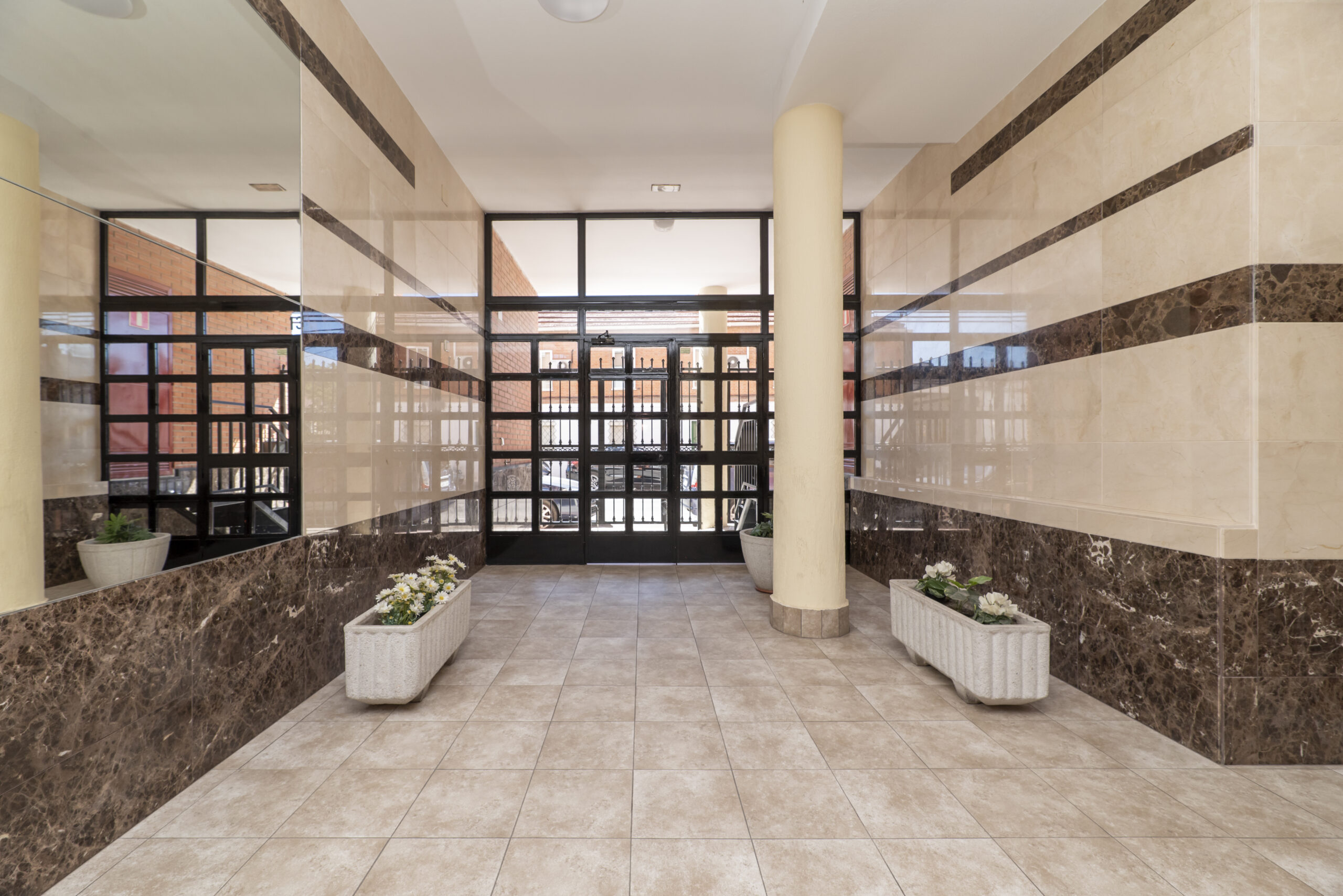Access portal to a residential building with white flower pots, a large black metal gate, walls covered in marble and mirrors